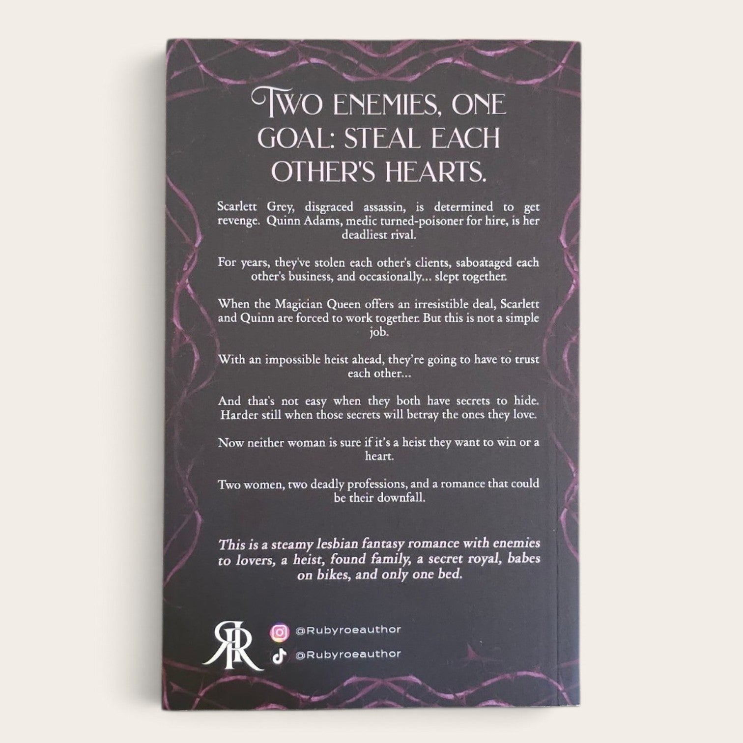 A book with a dark purple and black cover, featuring the text 'Two Enemies, One Goal: Steal Each Other Hearts' and a long poem in white text. The bottom includes social handles '@rubycoeauthor' and a monogram logo, photographed against a plain light background.
