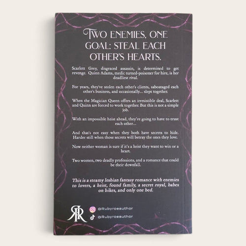 A book with a dark purple and black cover, featuring the text 'Two Enemies, One Goal: Steal Each Other Hearts' and a long poem in white text. The bottom includes social handles '@rubycoeauthor' and a monogram logo, photographed against a plain light background.