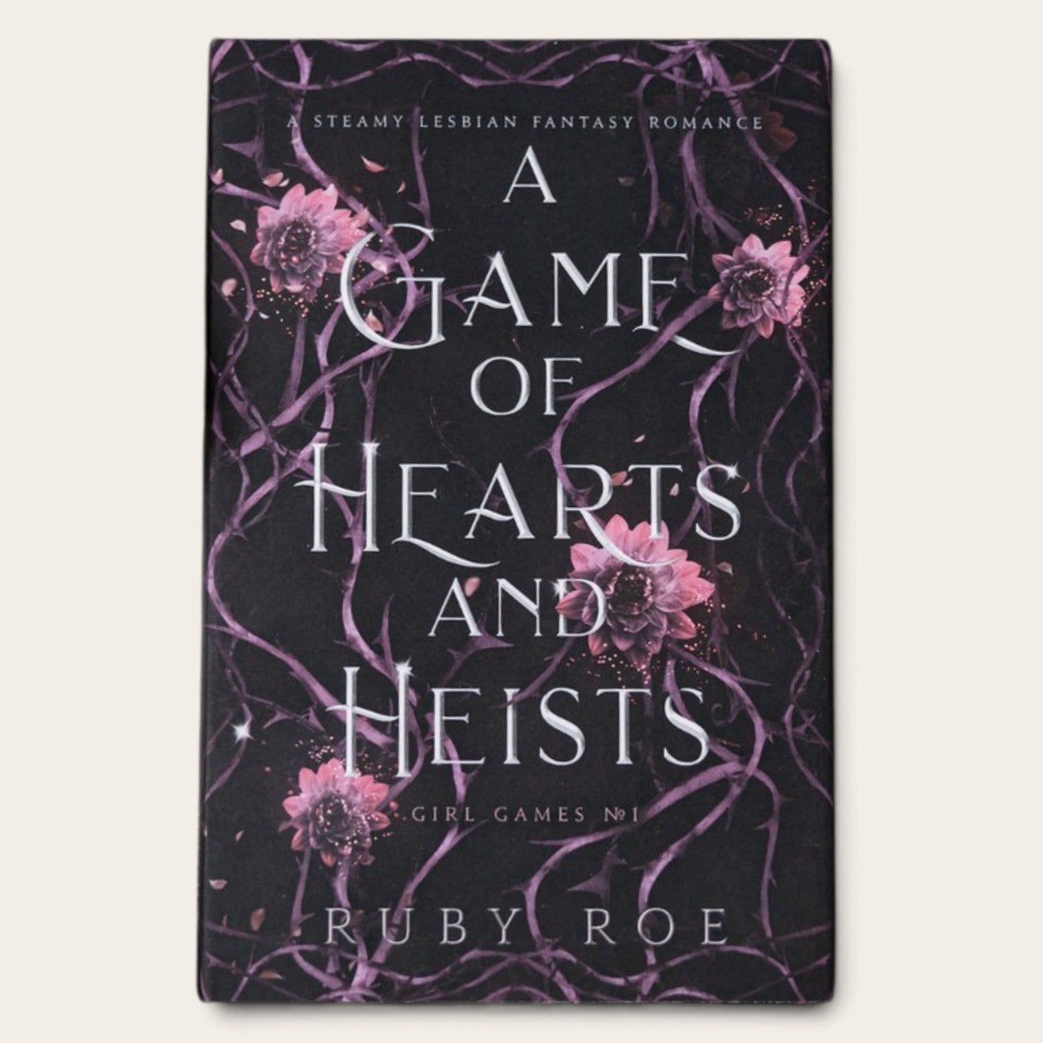 The book cover of 'A Game of Hearts and Heists' by Ruby Roe, featuring a dark background with purple vine patterns and pink flowers, displayed on a light beige background.