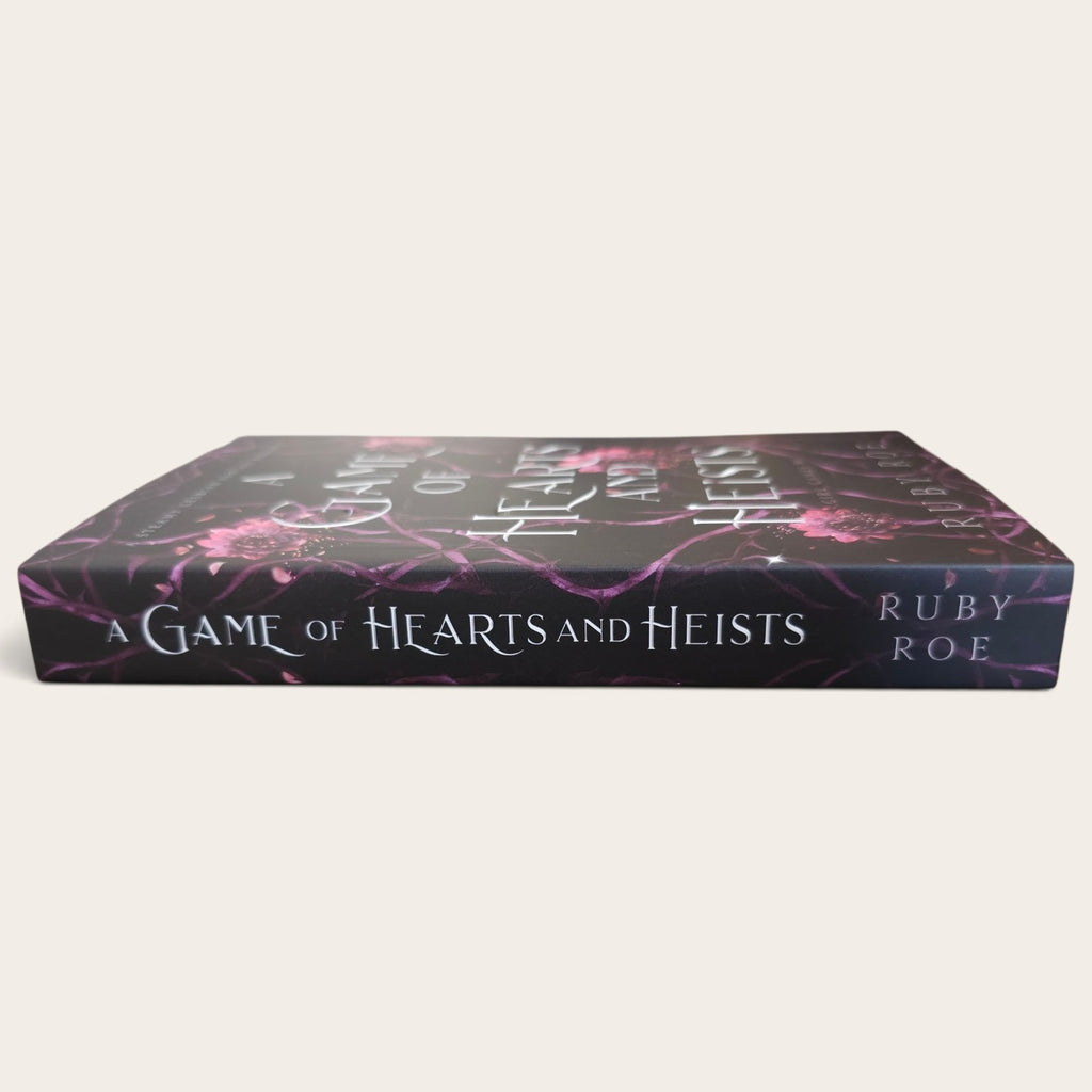 The book 'A Game of Hearts and Heists' by Ruby Roe, shown from a low angle against a light beige background. The book features a dark purple and black cover with pink floral patterns and white text.