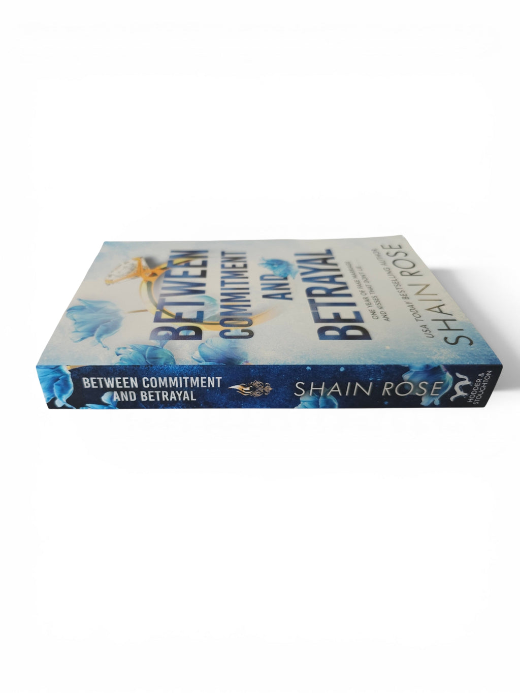 The book 'Between Commitment and Betrayal' by Shain Rose, shown at an angle on a clean white background. The cover features a light blue and white floral design with dark blue text and accents.