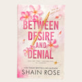 Book cover for 'Between Desire and Denial' by Shain Rose, featuring pink plumeria flowers and a gold pen on a textured light pink and white background.