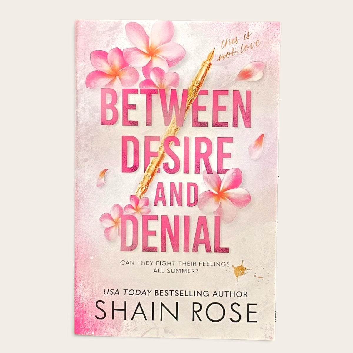 Book cover for 'Between Desire and Denial' by Shain Rose, featuring pink plumeria flowers and a gold pen on a textured light pink and white background.