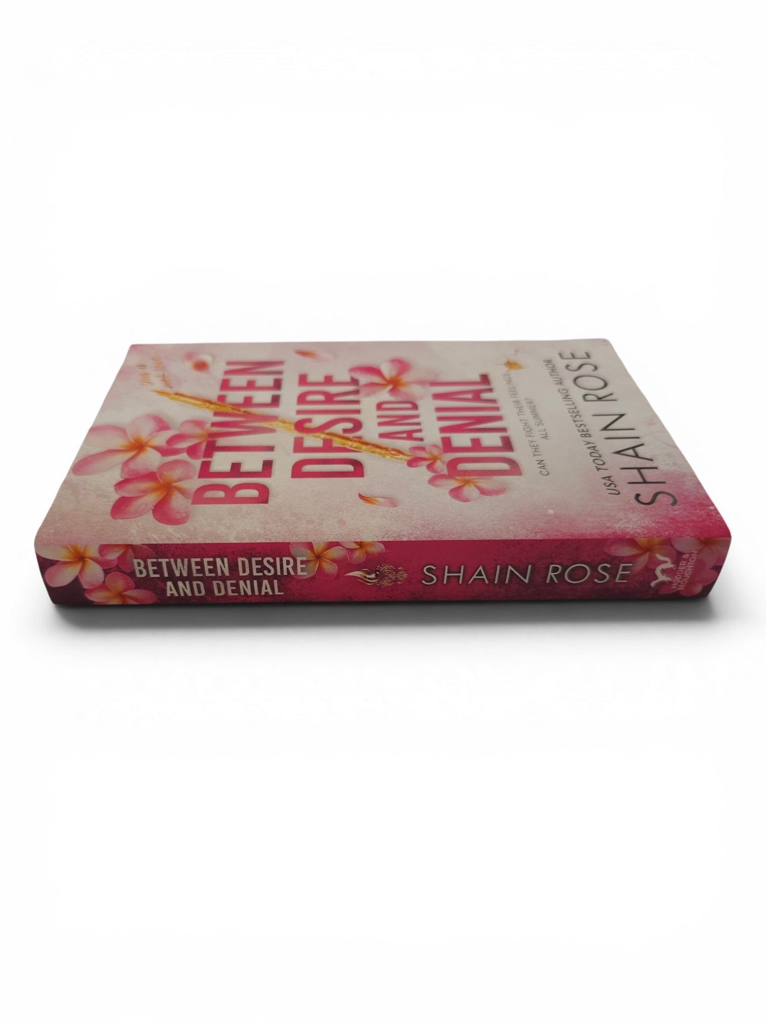 A paperback copy of the book 'Between Desire and Denial' by Shain Rose, featuring a pink and white cover with floral illustrations and a gold diagonal line.