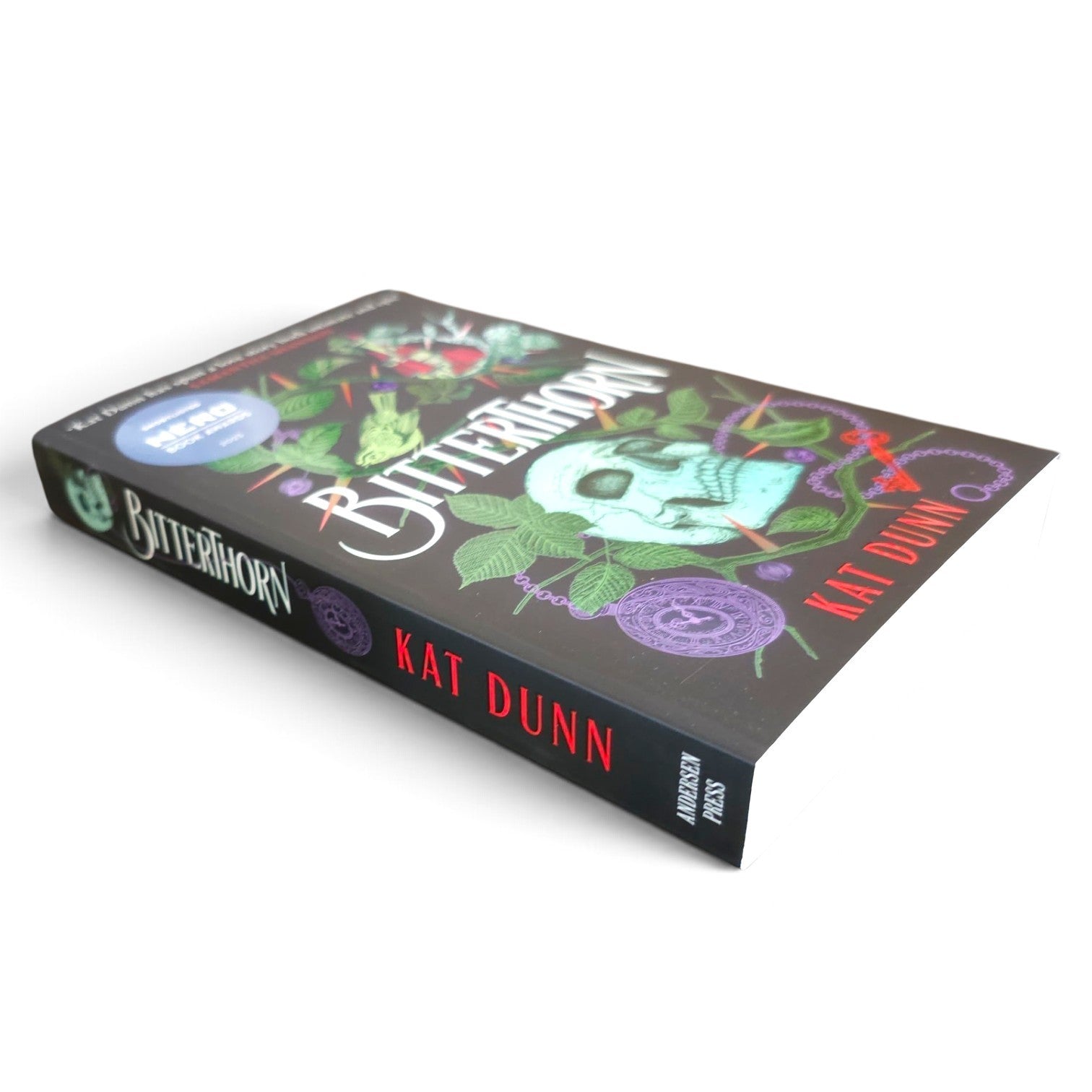 A book titled "Bitterhorn" by Kat Dunn, shown at an angle on a plain white background. The cover is dark gray with a skull, green leaves, purple circular motifs, and red title text.