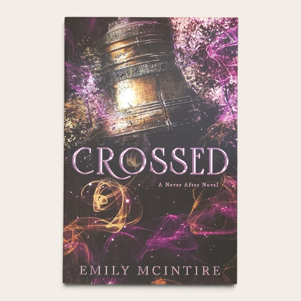 Book cover for 'Crossed' by Emily McIntire, featuring a large bronze bell against a dark background with purple and gold glowing swirls and the title in large metallic letters.