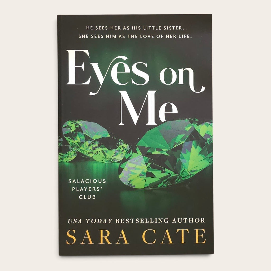 Book cover for 'Eyes on Me' by Sara Cate, featuring a dark background with large white title text, green faceted gemstones, and gold author text, photographed against a light neutral background.