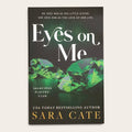 Book cover for 'Eyes on Me' by Sara Cate, featuring a dark background with large white title text, green faceted gemstones, and gold author text, photographed against a light neutral background.