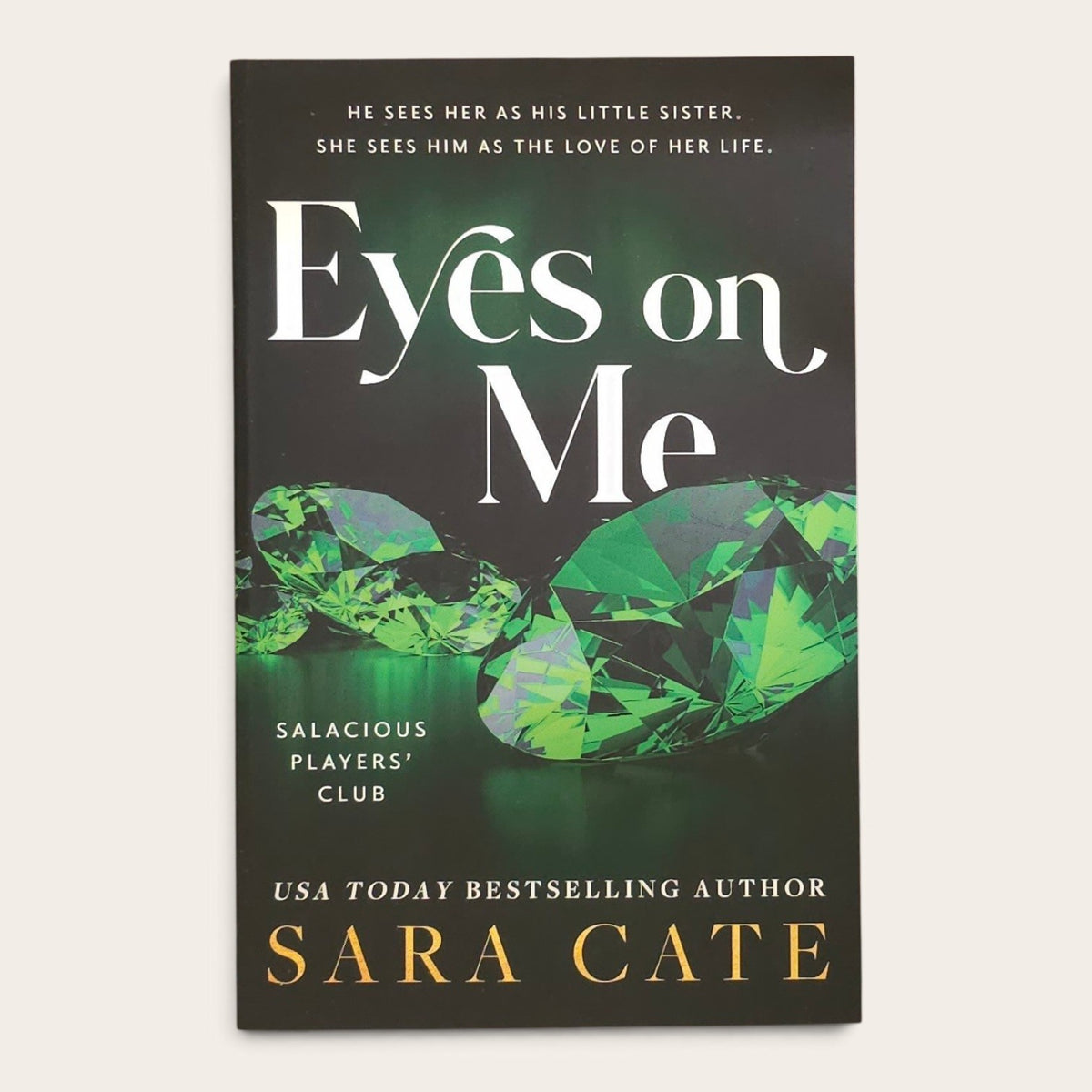 Book cover for 'Eyes on Me' by Sara Cate, featuring a dark background with large white title text, green faceted gemstones, and gold author text, photographed against a light neutral background.