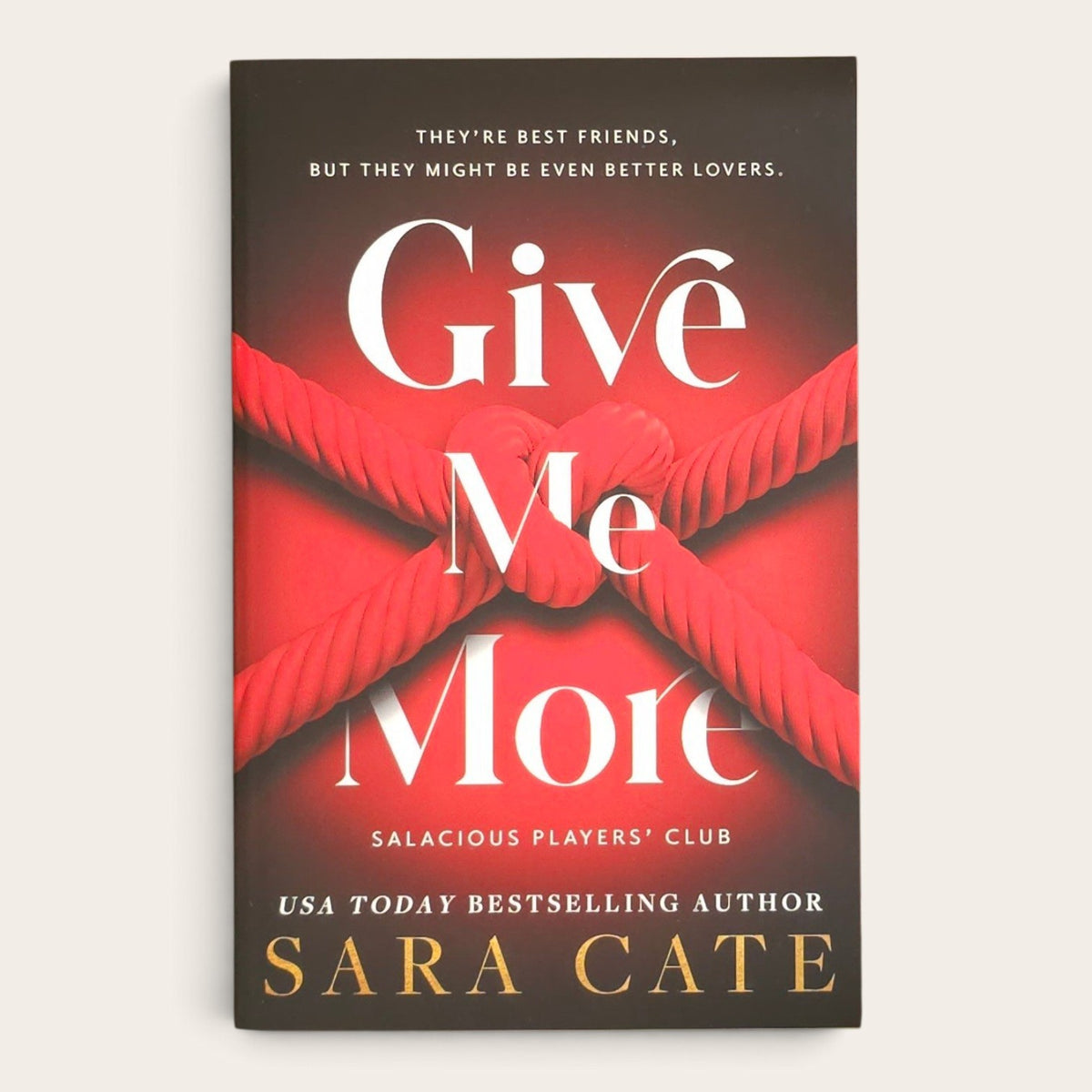 The book cover of 'Give Me More' by Sara Cate, featuring a dark brown and red background with a red rope knot design and white and gold text, shown on a light beige background.