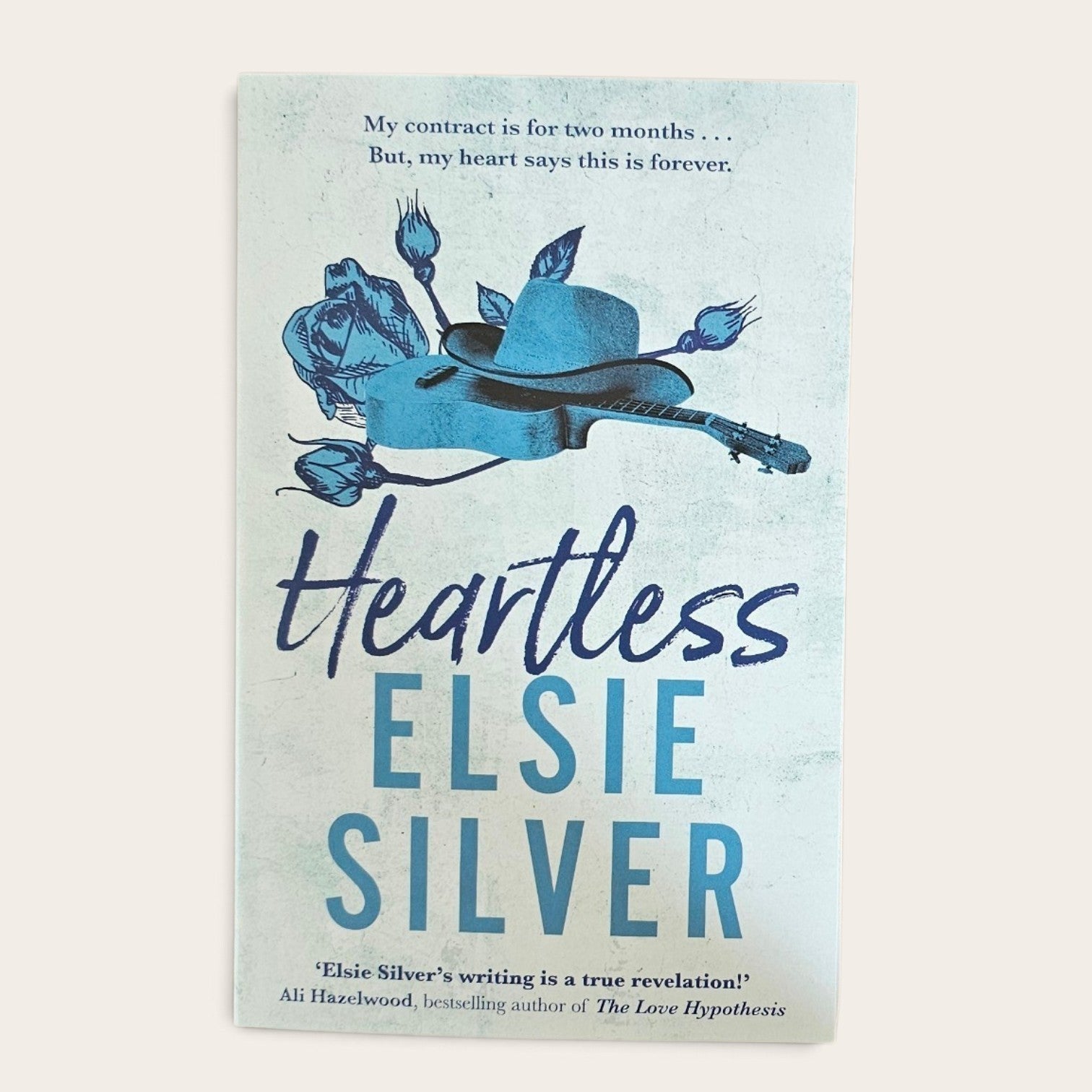 A book cover for 'Heartless' by Elsie Silver, featuring a light blue textured background with a blue illustration of a guitar, roses, and a hat, set against a warm off-white background.
