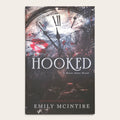 Book cover for 'Hooked: A Never After Novel' by Emily McIntire, featuring a dark clock face with Roman numerals, red lipstick marks, and distressed splatter artwork on a light beige background.