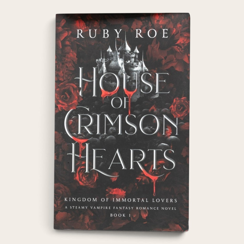 Book cover for 'House of Crimson Hearts' by Ruby Roe, featuring a gothic castle silhouette over a dark background with red roses and red blood-like drips, shown on a light beige background.