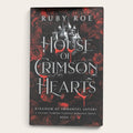 Book cover for 'House of Crimson Hearts' by Ruby Roe, featuring a gothic castle silhouette over a dark background with red roses and red blood-like drips, shown on a light beige background.