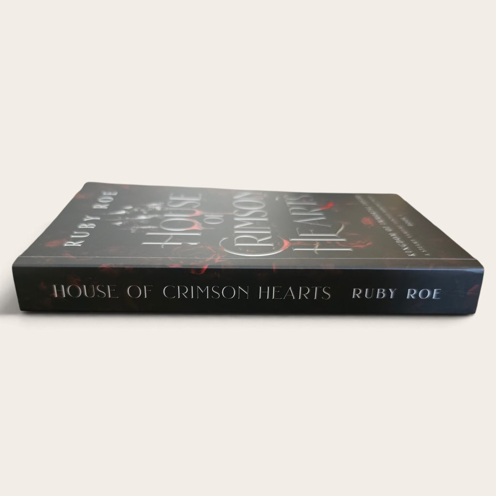 A paperback book titled 'House of Crimson Hearts' by Ruby Roe, shown at an angle on a light beige background. The cover is dark gray with white gothic-style lettering and red accents.