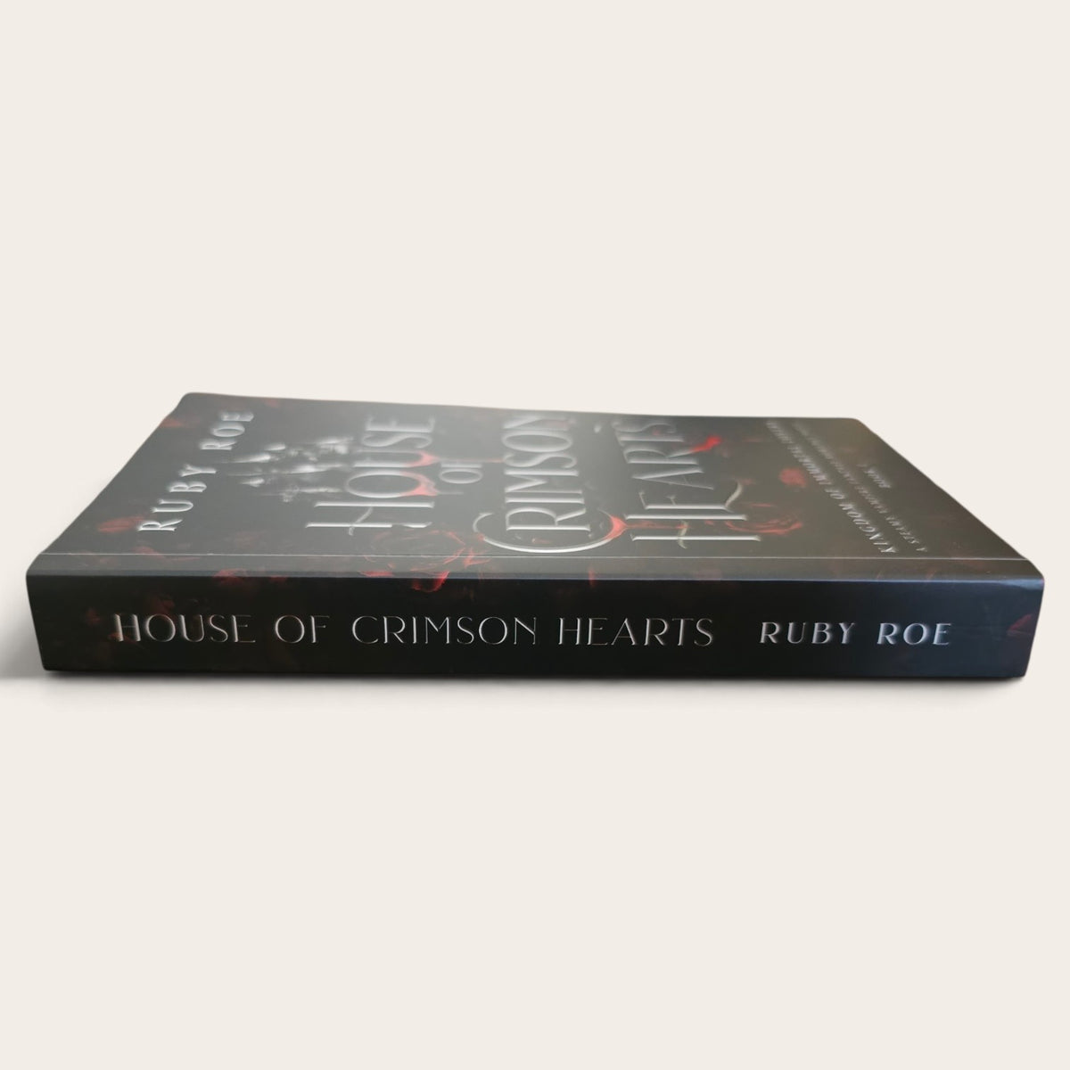 A paperback book titled 'House of Crimson Hearts' by Ruby Roe, shown at an angle on a light beige background. The cover is dark gray with white gothic-style lettering and red accents.