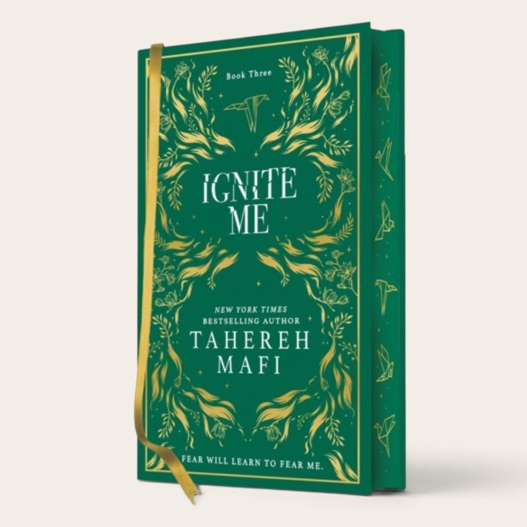 The book 'Ignite Me' by Tahereh Mafi, shown as a hardcover edition with a deep green cover, gold botanical illustrations, and a gold ribbon bookmark, set against a plain off-white background.