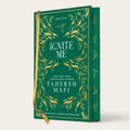 The book 'Ignite Me' by Tahereh Mafi, shown as a hardcover edition with a deep green cover, gold botanical illustrations, and a gold ribbon bookmark, set against a plain off-white background.