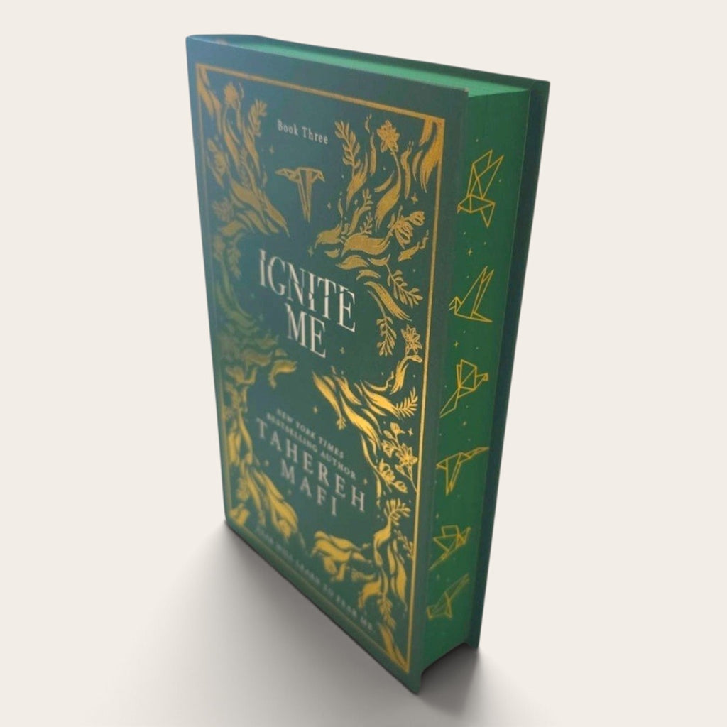 A hardcover book titled 'Ignite Me' by Tahereh Mafri, featuring a dark green cover with ornate gold botanical and bird illustrations, shown at an angle on a light beige background.