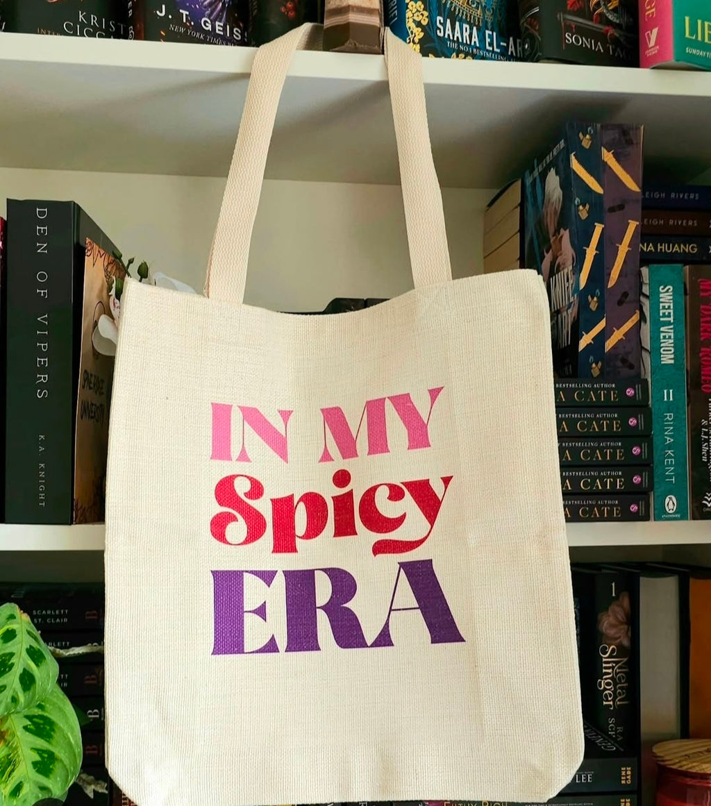 A natural canvas tote bag hanging on a bookshelf, printed with the text 'IN MY Spicy ERA' in pink, red, and purple lettering.