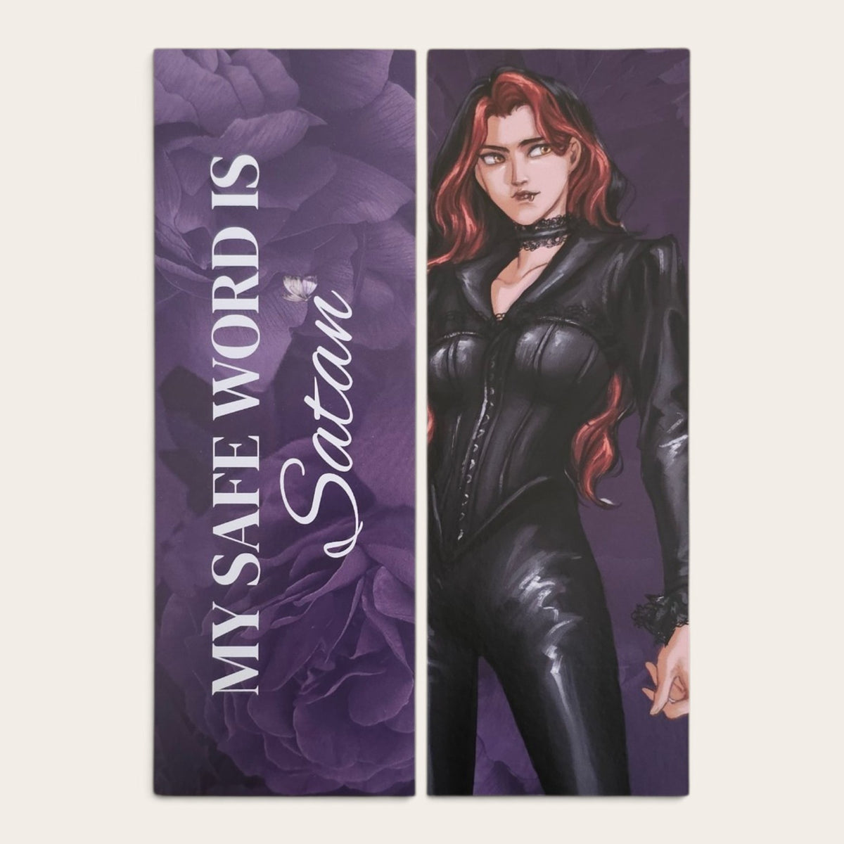 a bookmark showing front and back featuring a stylized illustration of a woman with red hair in a black catsuit against a purple background, alongside the text 'MY SAFE WORD IS Satan' on a purple rose backdrop.