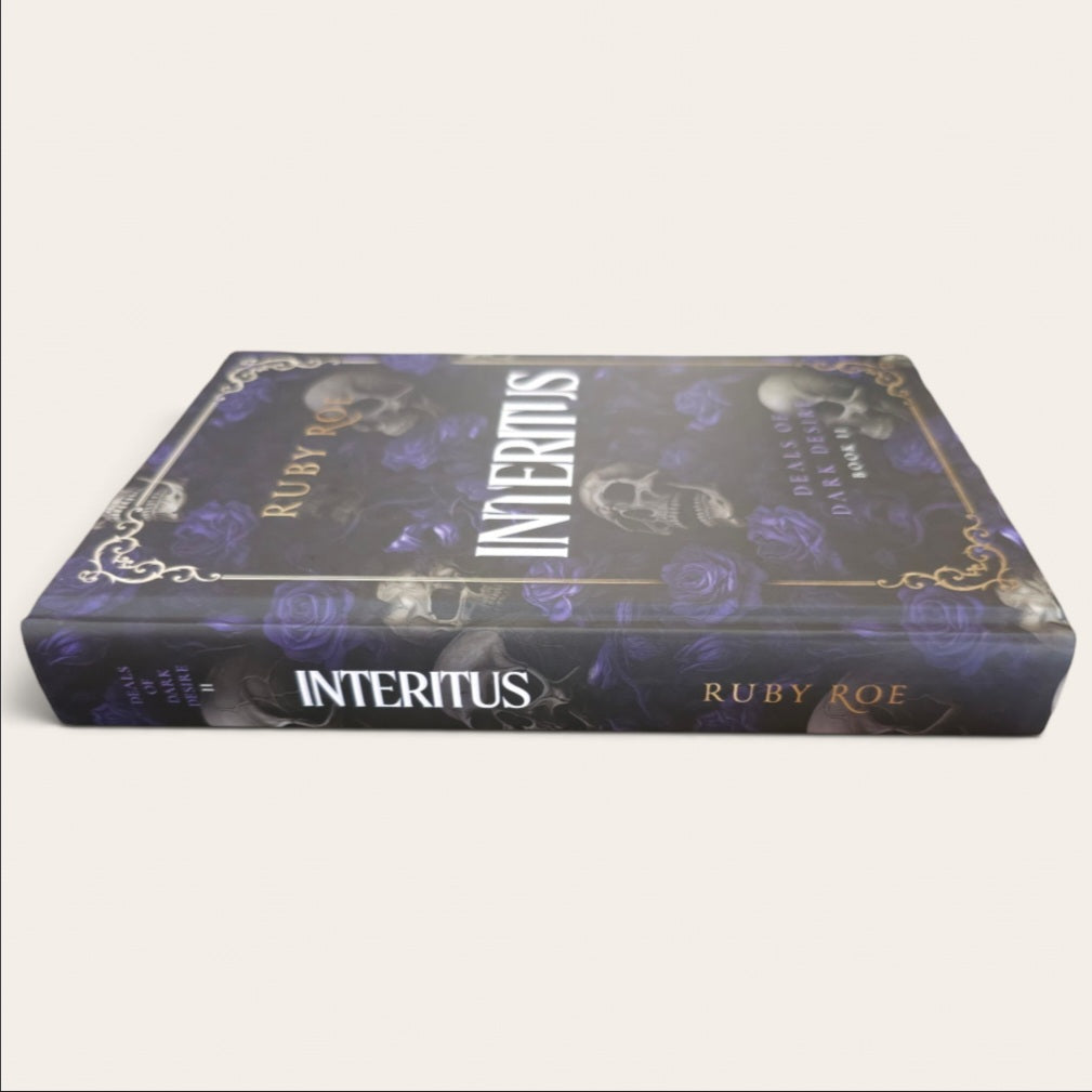 The book 'Interitus' by Ruby Roe, featuring a dark purple and black cover with gold ornamental borders, white title text, and skull illustrations, shown on a light beige background.