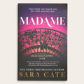 The book cover of 'Madame' by Sara Cate, featuring a dark purple and black background with a pink and red patterned fan illustration and gold and white text.