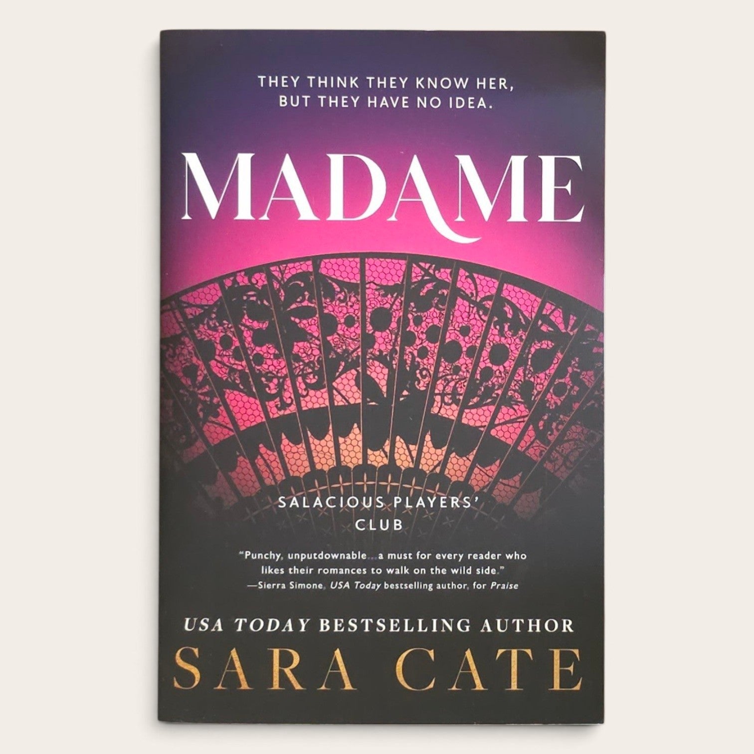 The book cover of 'Madame' by Sara Cate, featuring a dark purple and black background with a pink and red patterned fan illustration and gold and white text.