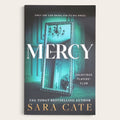 Book cover for 'Mercy' by Sara Cate, featuring a dark teal and black background with a large ornate mirror reflecting an interior scene. The title 'MERCY' is printed in large white letters, with the author name 'SARA CATE' in gold at the bottom.