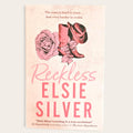 Book cover for 'Reckless' by Elsie Silver, featuring a pink and beige western-themed design with a rose, cowboy boots, and hat on a light cream background.