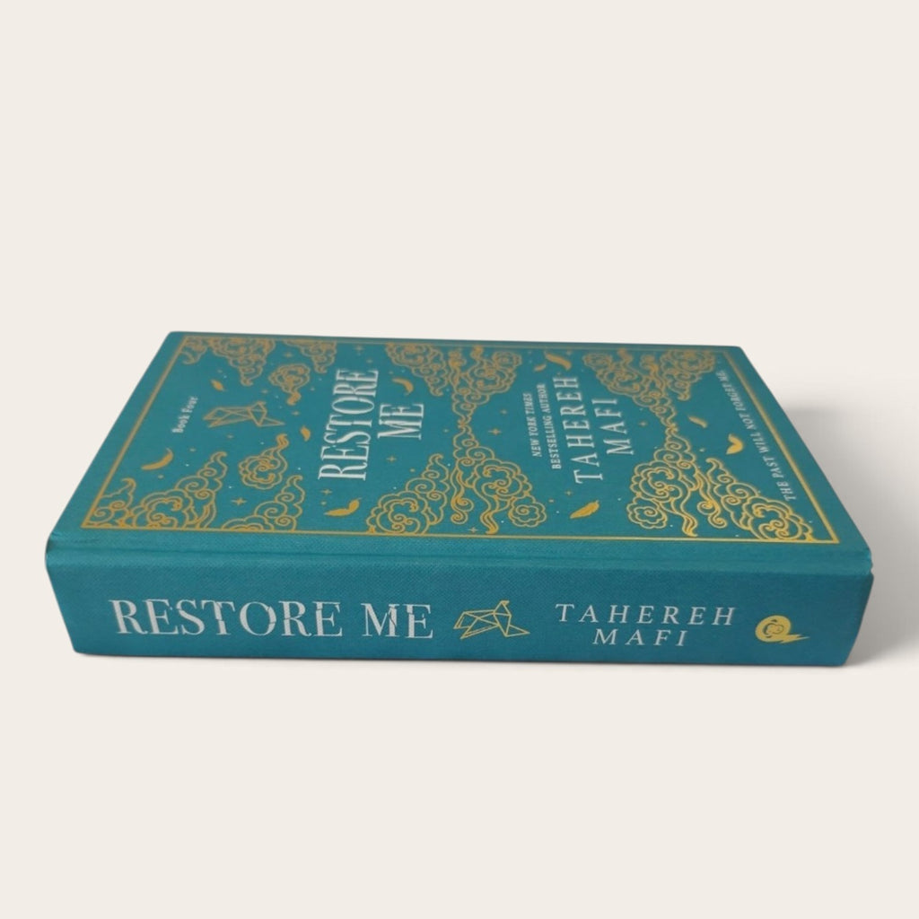 A hardcover book titled 'Restore Me' by Tahereh Mafi, featuring a teal-blue cover with ornate gold foil patterns and lettering, photographed on a clean off-white background.