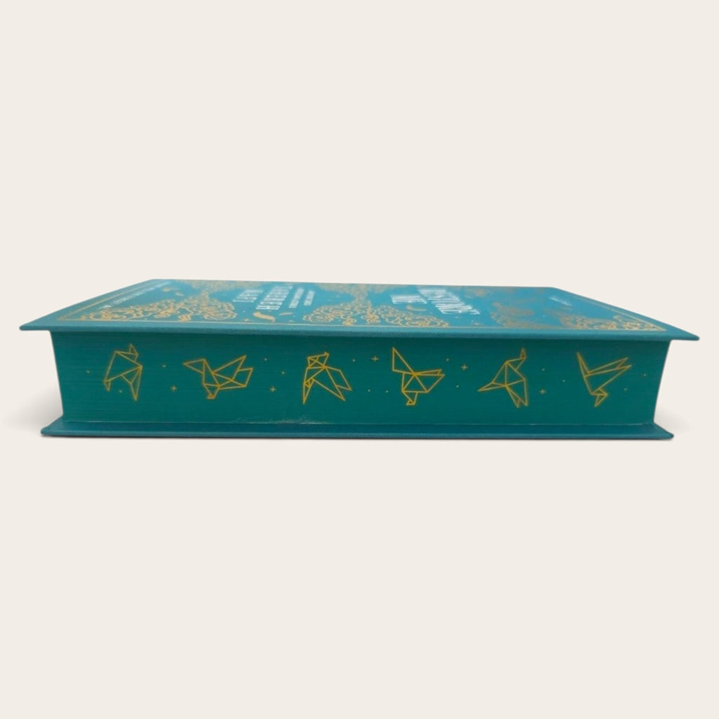A teal book-shaped decorative box with gold foil illustrations of origami cranes and stars, shown on a plain light beige background.