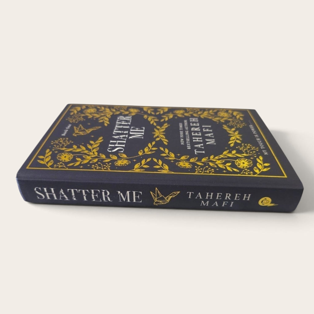 A hardcover book titled 'Shatter Me' by Tahereh Mafi, featuring a dark navy blue cover with intricate gold floral illustrations and gold lettering on the spine, set against a plain off-white background.