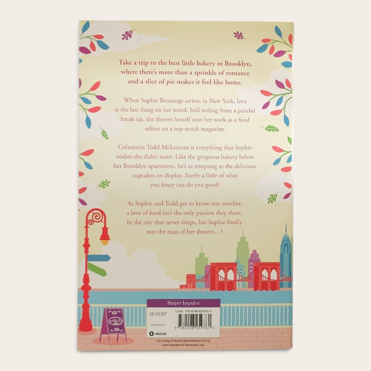 The back cover of a book titled 'The Little Brooklyn Bakery' by Julie Caplin, featuring a pastel illustration of a Brooklyn skyline with red bridges, a lamppost, and colorful leafy borders, on a light cream background.