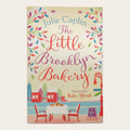 The front cover of the book 'The Little Brooklyn Bakery' by Julie Caplin, featuring a cream background decorated with colorful floral borders and an illustration of a bakery scene with tables, chairs, and a red bridge.