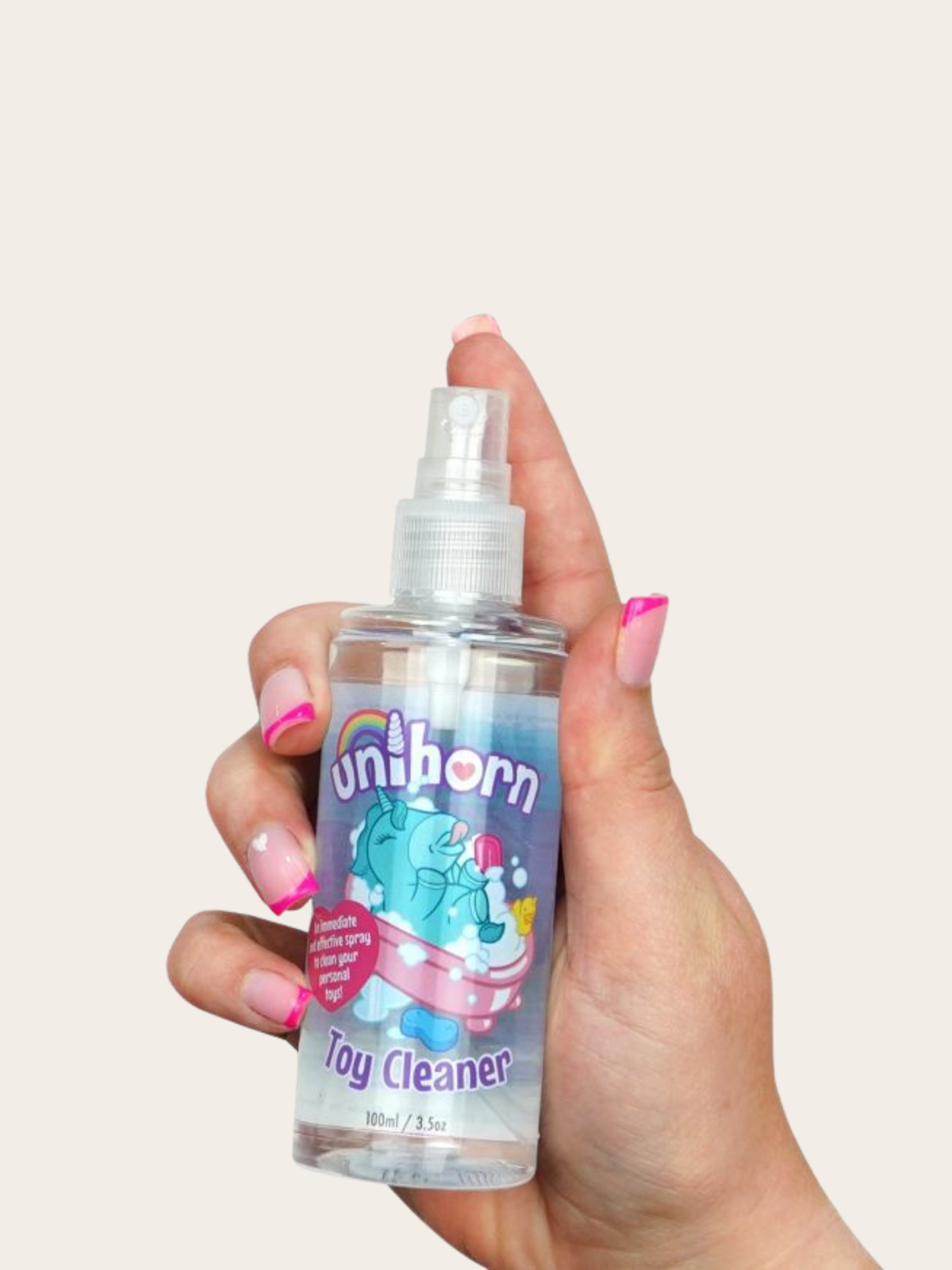 A hand with pink manicured nails holds a small clear plastic spray bottle labeled 'Unihorn Toy Cleaner' against a solid light beige background.
