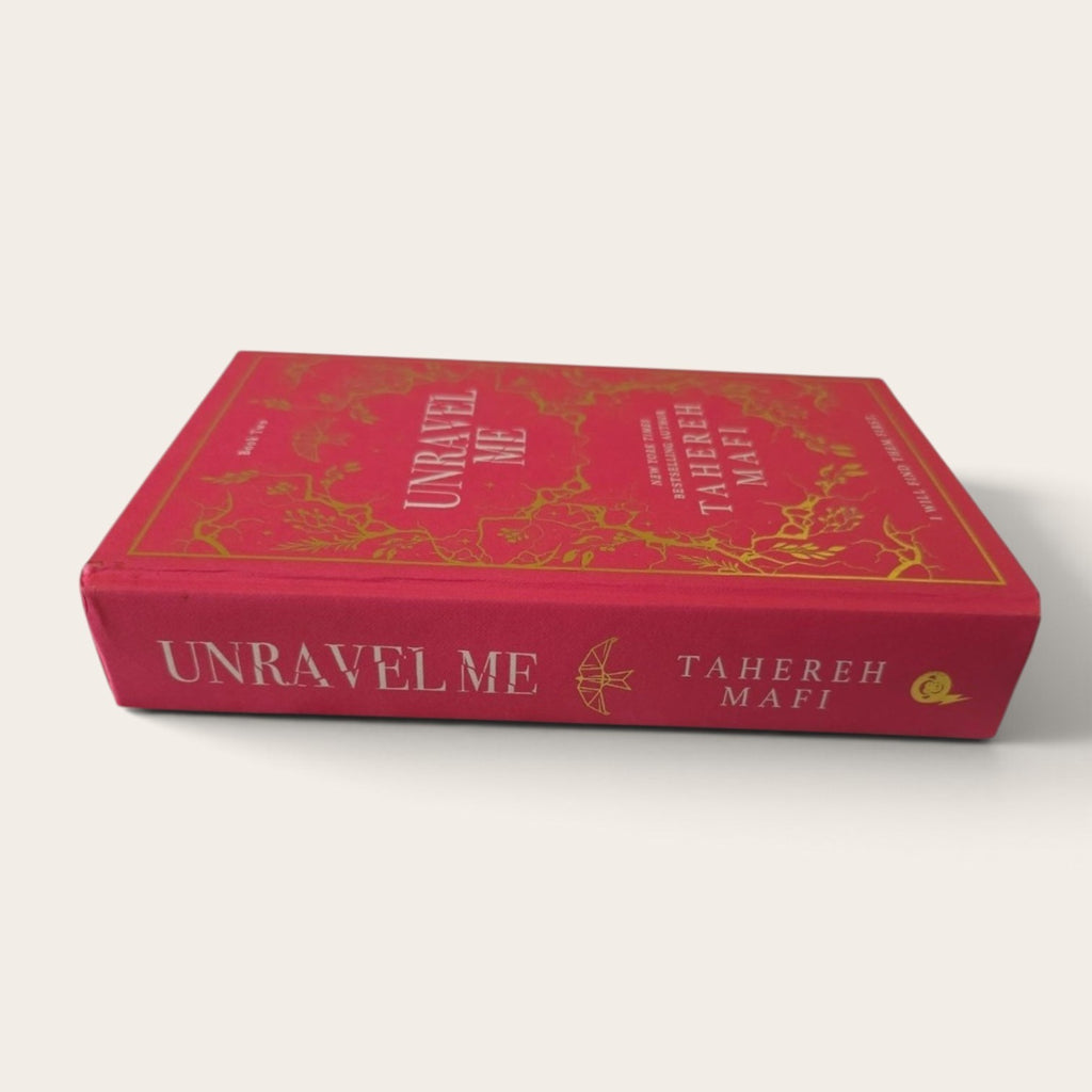 A hardcover book titled 'Unravel Me' by Tahereh Mafi, featuring a vibrant red cover with elegant gold foil detailing and white lettering on the spine, set against a clean off-white background.